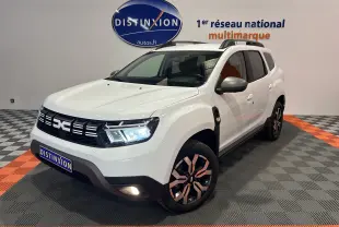 Dacia Duster BluedCi 115 Journey 4x4 blanc en 3/4 avant droit avec jantes noires et grises, phares allumés en showroom.