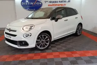 Fiat 500X blanc à toit noir en 3/4 avant droit, avec jantes sport et feux allumés dans un showroom.
