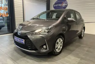 Toyota Yaris hybride gris foncé vue 3/4 avant droit en intérieur avec calandre noir laqué et phares allumés.