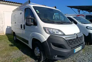 Vue 3/4 avant d'un utilitaire blanc Citroën Jumper L1H1 avec galerie de toit et pare-chocs noir sur un sol gravillonné.