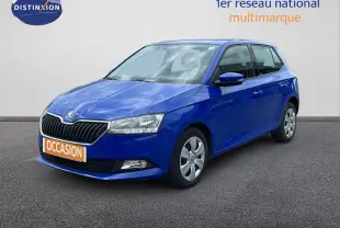 Skoda Fabia 1.0 TSI bleu énergie vue 3/4 avant, garée devant un garage avec façade vitrée et enseignes rouges.