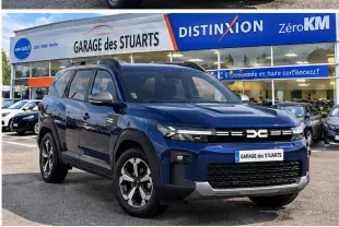 Dacia Bigster 2026 bleu indigo en 3/4 avant droit, avec jantes distinctives et toit noir, stationné devant un garage.