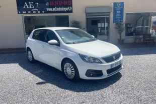 PEUGEOT 308 blanc vue 3/4 avant droit, garée devant un bâtiment avec panneaux publicitaires visibles.