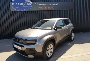 Jeep Avenger eHYBRID gris en 3/4 avant droit, avec calandre distinctive et jantes argentées, stationné devant un mur noir.