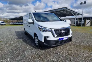 Nissan Primastar blanc en vue 3/4 avant droit sur parking extérieur avec abri métallique en arrière-plan.