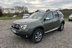 Dacia Duster 1.5 DCI 110 vert foncé en 3/4 avant droit, avec barres de toit et jantes alliage visibles.
