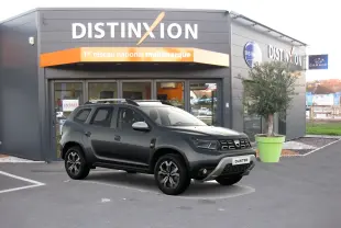 Vue 3/4 avant d'un Dacia Duster gris 2021 garé devant un showroom Distinxion avec ses jantes noires distinctives.