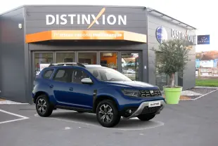 Dacia Duster bleu 2021 en 3/4 avant droit, avec jantes noires et toit argenté devant un showroom Distinxion.