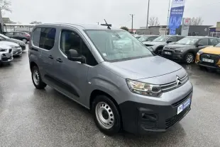 Citroën Berlingo van gris métal vu de trois quarts avant droit sur parking humide avec phares allumés.