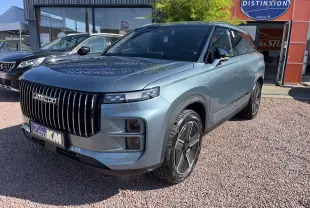 Vue 3/4 avant d'un JAECOO J7 gris olive avec calandre verticale noire et jantes modernes, stationné devant un showroom.