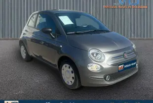 Vue 3/4 avant d'une FIAT 500 gris métallisé 2023 avec phares allumés sur fond urbain neutre.
