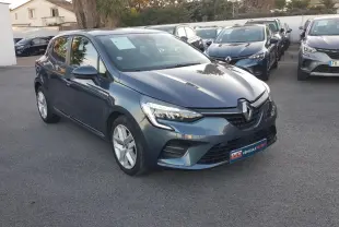 Renault Clio gris en vue 3/4 avant droit, avec phares allumés et jantes alliage visibles sur parking.