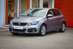 Peugeot 308 gris Artense vue 3/4 avant droit, avec calandre noire et jantes alliage, stationnée devant un bâtiment orange.