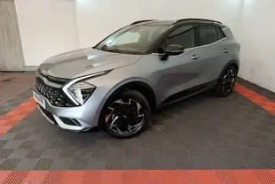 KIA Sportage gris clair en 3/4 avant droit, avec jantes noires et toit noir dans un garage éclairé.