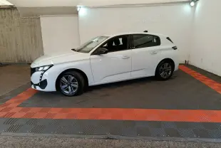 Peugeot 308 blanche vue de profil côté gauche, dans un espace fermé avec sol gris et bandes rouges.