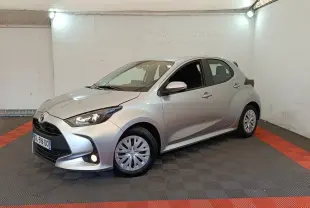 Toyota Yaris hybride gris clair vue 3/4 avant droit dans un garage avec feux allumés et jantes standard.