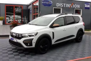Vue 3/4 avant droit du Dacia Jogger blanc avec détails noirs et rétroviseurs marron, stationné devant un bâtiment commercial.