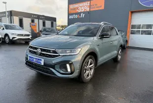 Volkswagen T-Roc bleu pétrole métallisé en 3/4 avant droit sur parking devant concession avec panneau promotionnel.