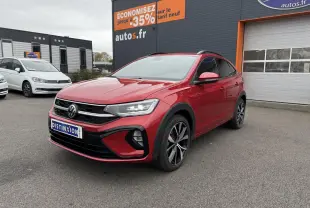 Volkswagen Taigo Rouge Roi en 3/4 avant droit, jantes noires brillantes, feux LED allumés, devant un garage autos.fr.