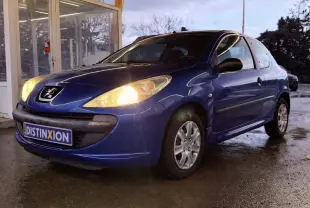 Peugeot 206+ bleu vue 3/4 avant droit avec phares allumés, garée devant un bâtiment vitré.
