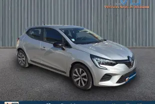 Renault Clio gris vue 3/4 avant droit sur sol bitumé devant un mur métallique, jantes alliage bicolore visibles.