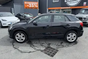 Audi Q2 noire vue de profil côté gauche, garée devant un garage avec panneaux publicitaires visibles.