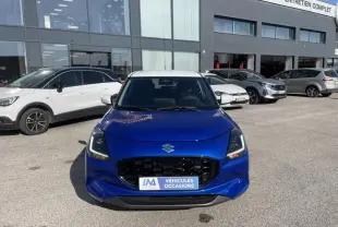 Suzuki Swift 1.2 Dualjet Hybrid bleu métal vue de face, calandre noire et toit blanc visible, devant un garage.