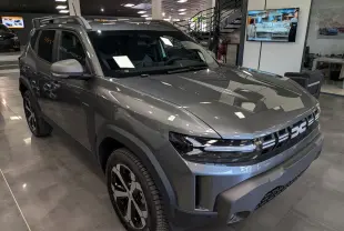 Vue 3/4 avant du Dacia Duster Hybrid-G 150 CH 4x4 gris schiste, avec calandre moderne et jantes bi-ton en showroom.