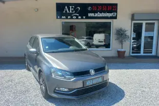 Volkswagen Polo gris vue 3/4 avant droit, avec calandre noire et jantes alliage, stationnée devant un commerce.