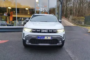 Vue frontale d’un Dacia Duster blanc glacier 2025 avec calandre noire et phares allumés devant un bâtiment vitré.