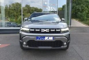 Vue avant d'un Dacia Duster 2026 gris schiste avec calandre noire et phares allumés, devant une vitrine de concession.