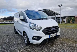 Ford Transit Custom blanc en vue 3/4 avant droit sur un sol gravillonné avec un ciel nuageux en arrière-plan.