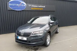 Skoda Karoq gris en 3/4 avant droit, SUV compact avec calandre noire et jantes argentées, stationné devant un mur noir.