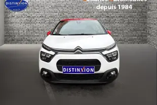 Vue frontale d'une Citroën C3 blanche avec toit et rétroviseurs rouges, sur fond de mur en briques grises.