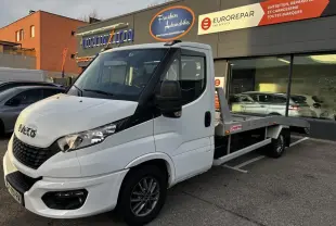 IVECO Daily blanc en 3/4 avant droit, plateau arrière visible, stationné devant un garage avec enseignes.