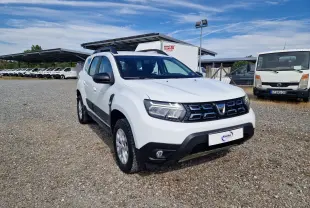 Dacia Duster 4x4 blanc vue 3/4 avant droit sur parking gravillonné avec calandre noire et jantes argentées.