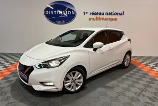 Vue 3/4 avant d'une Nissan Micra blanche 5 portes, en intérieur sur sol gris et orange, avec phares allumés.