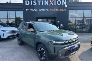 Vue 3/4 avant droit d'un Dacia Duster 2025 kaki dusty avec jantes alliage et barres de toit noires devant un showroom Distinxion.