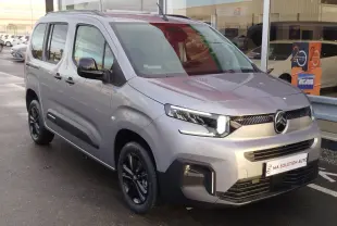 Vue 3/4 avant droit d'un Citroën Berlingo gris acier avec jantes noires et feux LED allumés, stationné devant un showroom.