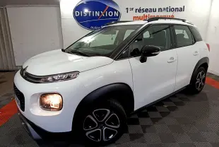 Vue 3/4 avant droite d'un Citroën C3 Aircross blanc avec phares allumés et barres de toit noires en intérieur showroom.