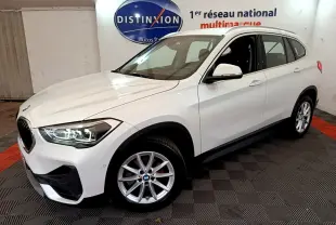 BMW X1 sDrive18i blanc vue 3/4 avant droit, phares LED allumés et jantes alliage visibles.