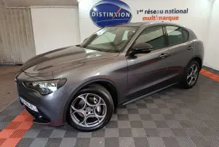 Alfa Romeo Stelvio gris foncé vu de 3/4 avant droit, avec jantes distinctives et phares LED allumés.