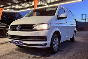Volkswagen Transporter L1H1 blanc vu en 3/4 avant droit, phare allumé, stationné sous un abri au crépuscule.