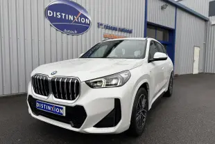 BMW X1 18D blanc minéral en vue 3/4 avant droit, avec calandre imposante et phares LED allumés.