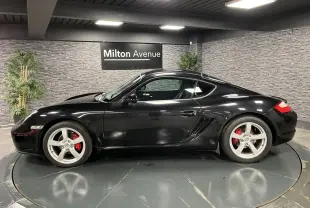 Profil côté gauche d'une Porsche Cayman noire 2008 avec jantes argentées et étriers de frein rouges en intérieur showroom.