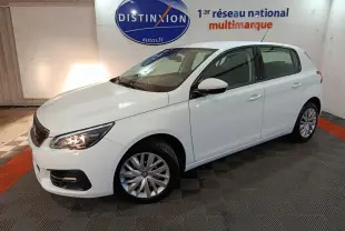 Peugeot 308 blanc vue 3/4 avant droit, phares allumés, stationnée en intérieur sur sol damier noir et rouge.