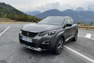 Peugeot 3008 GT Line gris Platinium vue 3/4 avant droit sur parking avec montagnes en arrière-plan.