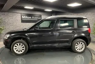 Profil côté gauche d'un Skoda Yeti noir 2015 avec jantes alliage 16'' et barres de toit argentées en intérieur showroom.