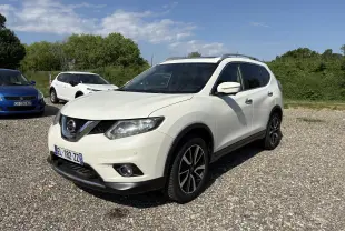 Nissan X-Trail 2016 blanc en 3/4 avant droit, avec jantes alliage et barres de toit visibles sur un sol gravillonné.