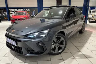 Vue 3/4 avant d'une CUPRA Leon gris Graphène avec jantes anthracite et signature lumineuse Tri-LED visible en intérieur showroom.